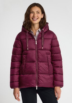 Giovane donna sorridente, indossa una giacca imbottita bordeaux con cappuccio e "Polo Club" coulisse, in piedi davanti a uno sfondo uniforme.