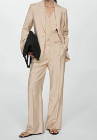 Costume beige à rayures fines avec pantalon à jambes larges et gilet ajusté, comprenant un blazer à simple boutonnage et un sac noir texturé.
