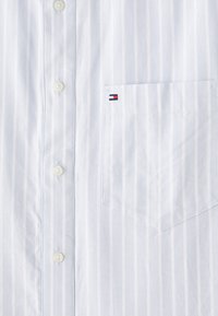 Chemise blanche à boutons avec des rayures verticales subtiles et une poche poitrine avec un petit écusson logo rouge, blanc et bleu.