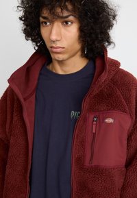 Jachetă din fleece burgund cu o suprafață texturată, buzunar frontal cu fermoar și accente roșii, purtată peste un tricou bleumarin cu text grafic.