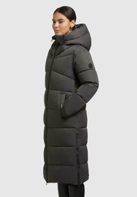 Piumino lungo in tessuto grigio scuro. Presenta un cappuccio, chiusura con zip e tasche laterali. Sezioni trapuntate orizzontali per l'isolamento.