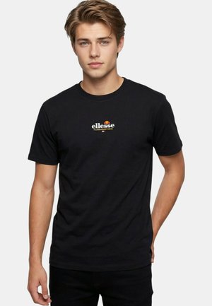 Jeune homme aux cheveux châtain clair portant un t-shirt noir Ellesse avec logo, posant devant un fond uni gris clair.