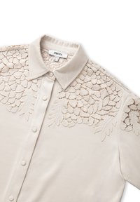 Lichtbeige blouse met een kraag, met ingewikkelde kantborduursels op de schouders en aan de voorkant. Soepele textuur, knoopsluiting.