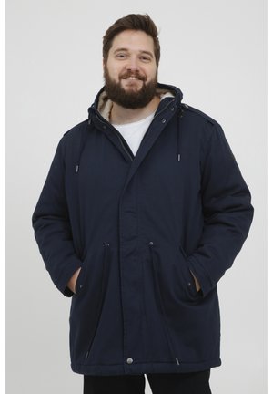 Parka de color azul marino con una suave capucha forrada de piel, cordones ajustables y bolsillos frontales. Cierre de botones y cremallera con un corte holgado.