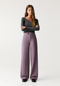 Pantalons longs taille haute en tissu lavande doux avec une fermeture à bouton et des plis à l'avant, associés à un haut gris ajusté à manches longues.