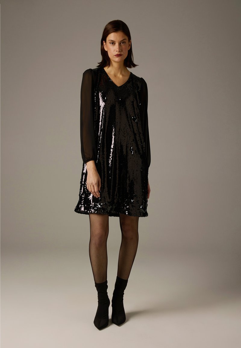 Schwarzes Paillettenkleid mit V-Ausschnitt, langen transparenten Ärmeln und ausgestelltem Saum. Kombiniert mit Netzstrumpfhosen und schwarzen Stiefeletten. Glatte Textur.