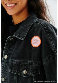 Czarna denimowa kurtka z okrągłą naszywką. Naszywka jest pomarańczowa z różowym napisem "Jak tam? PRZYJACIELE." Widoczne srebrne guziki i szwy.