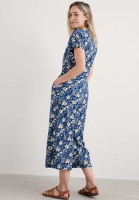 Kombinezon w kwiatowy wzór w kolorze granatowym z białymi i jasnoniebieskimi liśćmi, z krótkimi rękawami, bocznymi kieszeniami i luźnym krojem. Beżowe sandały w zestawie.