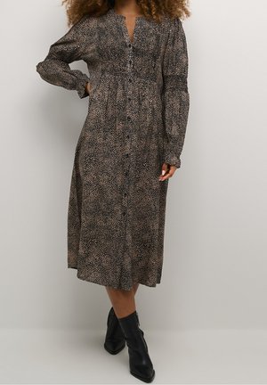 Femme portant une robe midi à manches longues boutonnée sur le devant avec un motif tacheté noir et beige, assortie à des bottines noires.