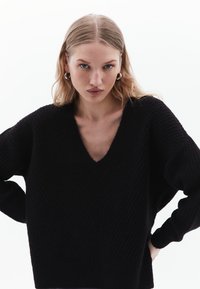 Maglione nero lavorato a maglia con scollo a V profondo, texture a coste e maniche ampie. Il tessuto appare morbido con una vestibilità comoda e dettagli sottili.