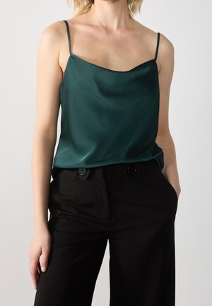 Femme portant un caraco en satin vert foncé à fines bretelles, rentré dans un pantalon noir taille haute avec des pattes de ceinture boutonnées.