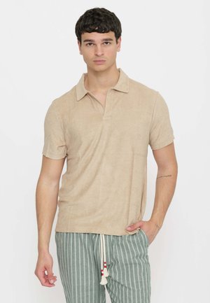 Giovane uomo con capelli corti e scuri indossa una polo beige a maniche corte e pantaloni verdi a righe con coulisse bianca, in piedi davanti a uno sfondo bianco.