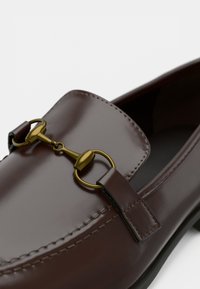 Mocassins en cuir marron à enfiler, dotés d'un mors accent doré et de détails de couture le long des bords. Texture lisse et forme arrondie.