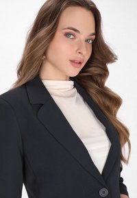 Czarna marynarka z klapkami, w połączeniu z kremową bluzką o stójce. Charakteryzuje się gładkimi fakturami i detalem w postaci guzików na marynarce.