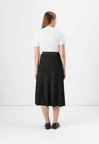 Una mujer está de espaldas al espectador, vistiendo una camiseta blanca y una falda midi negra y brillante, sobre un fondo blanco.