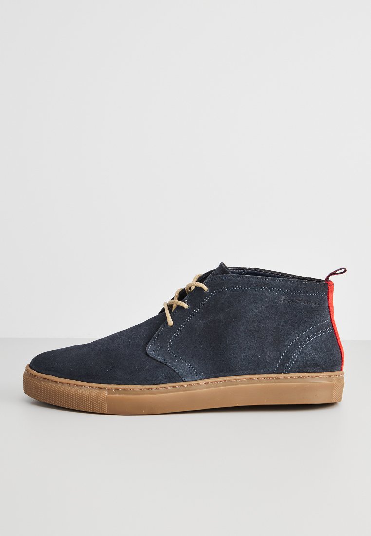 Blauwe suède enkelboots met een tan rubberen zool, beige veters en een rode hielaccent. Kenmerkt zich door een gestikt ontwerp en een gladde textuur.