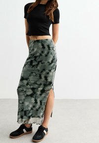 Frau in einem schwarzen, kurzen T-Shirt, einem grün-schwarzen Batik-Maxirock mit seitlichem Schlitz und schwarzer Sneakers mit weißen Streifen.