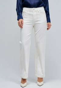 Femme portant un jean taille haute blanc à jambes droites, une chemise cropped bleu marine et des escarpins blancs à bout pointu, se tenant devant un fond uni.