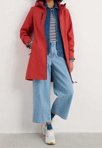 Rød vandtæt frakke med hætte, lagdelt over en denimjakke og en stribet top. Vide blå jeans og hvide sneakers fuldender outfittet.