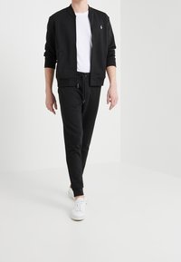 Man som går och bär en svart zip-up jacka, vit t-shirt, svarta dragbandsbyxor och vita sneakers mot en vit bakgrund.