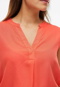Top senza maniche color corallo arancione con scollatura a V profonda e delicate pieghe. Realizzato in tessuto leggero con una texture liscia.