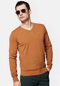 Brązowy sweter w serek z dzianiny, z długimi rękawami i dopasowanym dołem. Noszony z zielonymi spodniami i okularami aviator.