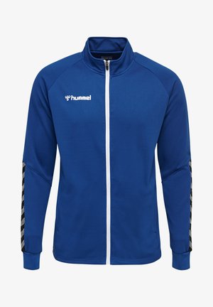 Blå atletisk jakke med tekstureret overflade, hvid lynlås og sort/sølv chevron-detaljer på ærmerne. Logo på venstre bryst.
