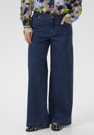 Dunkelblaue, weit geschnittene Jeans mit seitlichen Knopfdetails, aus Denim gefertigt. Kombiniert mit einem farbenfrohen, gemusterten Langarmhemd und schwarzen, mit Nieten besetzten Schuhen.