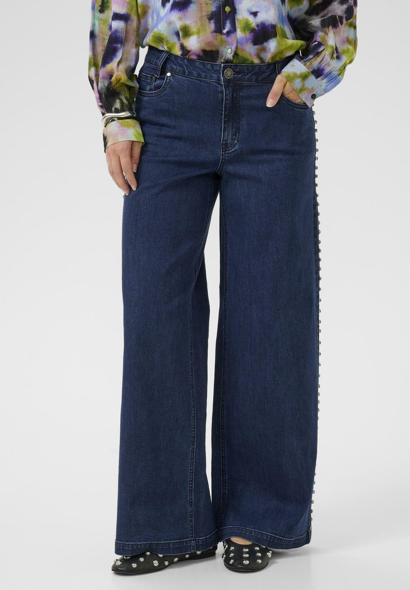 Temno modri široki jeans s stranskimi gumbi, narejeni iz denima. Kombinirani z barvno, vzorčasto dolgotrajno srajco in črnimi čevlji s studenci.