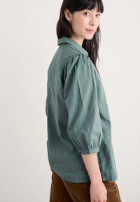 Bluza s pufastimi rukavima v tihem teal odtenku, z klasičnim ovratnikom, vertikalnimi črtami in nekoliko ohlapnim krojem. Narejena iz lahkega blaga.