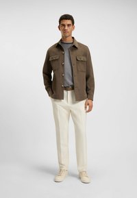 Men's brown jacket with large front pockets, gray crew-neck shirt underneath, cream trousers, and beige sneakers. Flat, smooth texture.