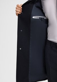 Zwarte jas met een gladde binnenvoering, voorzien van knopen aan de voorkant en een subtiele zakdetail aan de binnenkant. Standaard reversontwerp.