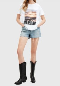 Bílý tričko s grafikou přírody a nápisem "ALLSAINTS", spárované s modrými šortkami a černými kovbojskými botami se vzorem plamenů.