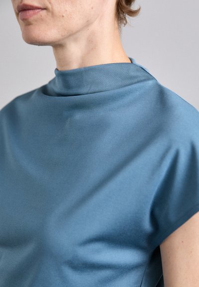 Blått, glatt stoff med høy hals og cap ermer. Har en drapert design med en subtil glans og minimalisme.