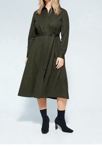 Robe longue à manches longues couleur vert foncé avec un bouton à l'avant, taille ceinturée et jupe fluide. Associée à des bottines noires à la cheville. Texture de tissu lisse.