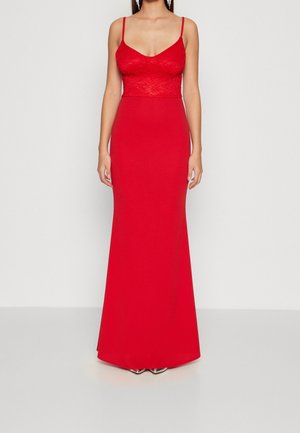 Woman wearing a sleeveless, floor-length red dress with lace bodice and thin straps, standing against a plain light background.