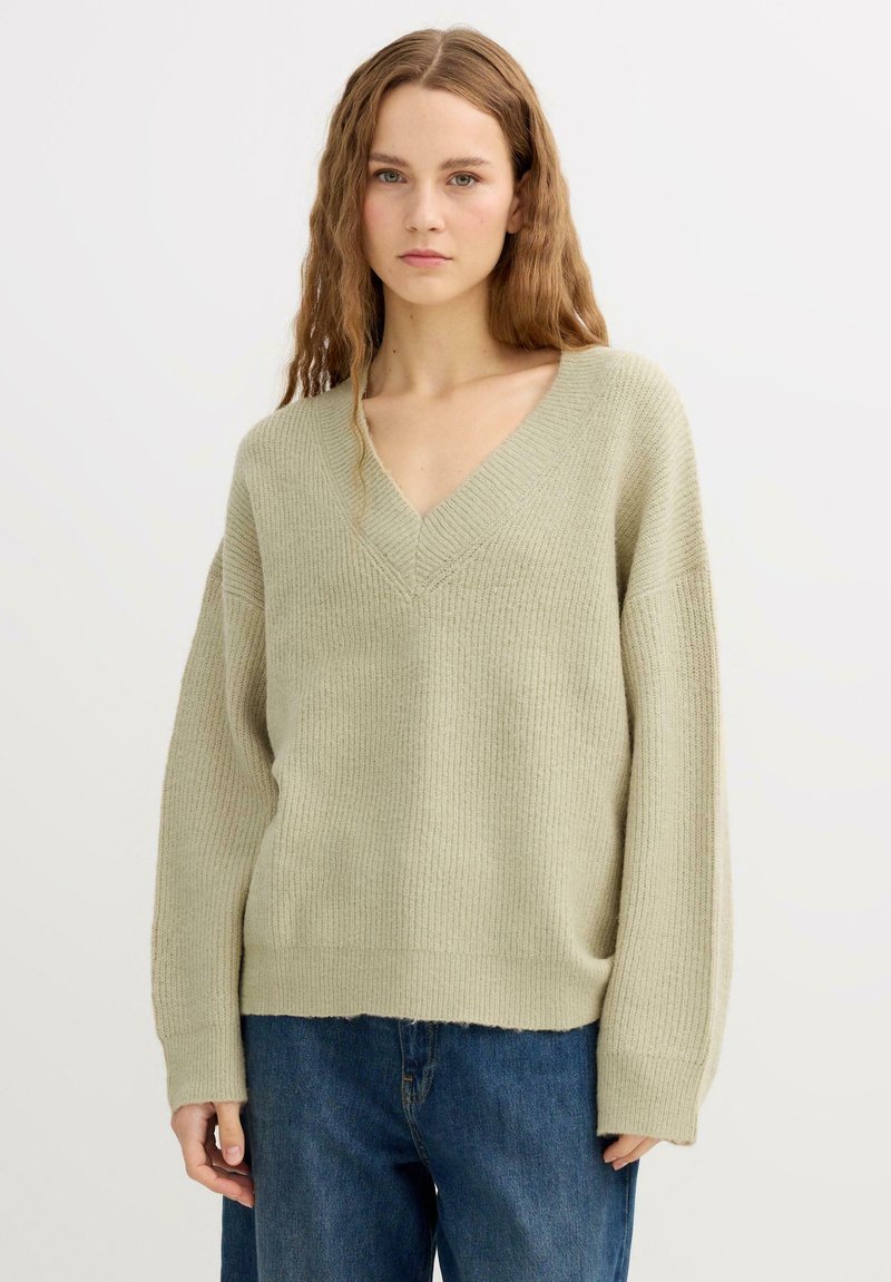 Suéter de punto acanalado verde claro con escote en V, hombros caídos y dobladillo recto. Combinado con vaqueros de mezclilla azul de pierna ancha.