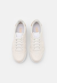 Un paio di sneakers bianche a basso top con accenti beige, lacci allacciati, viste dall'alto su uno sfondo grigio chiaro uniforme.