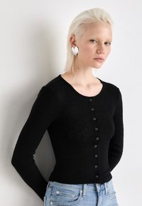 Fekete bordázott, hosszú ujjú felső kerek nyakkivágással és elülső gombos záródással, világoskék denim farmerral párosítva. Ezüst szív alakú fülbevalók.