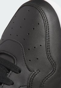 Zapatilla negra de cuero con detalles perforados, costuras texturizadas y cordones planos negros, que cuenta con una suela de goma blanca.