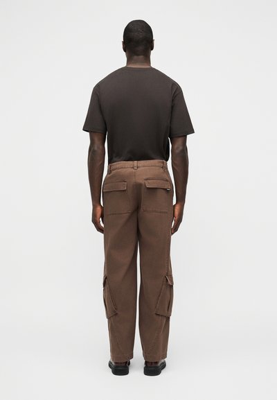 Άνδρας που φοράει μαύρο κοντομάνικο πουκάμισο και φαρδιά καφέ παντελόνια cargo με πίσω τσέπες με καπάκι, στέκεται με την πλάτη προς τα εμπρός σε απλό φωτεινό φόντο.