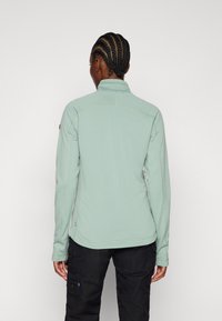 Hellgrüner Pullover mit langen Ärmeln aus strukturiertem Stoff, hohem Kragen und kontrastierenden Nähdetails. Getragen mit schwarzen Cargo-Hosen.