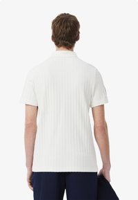 Vit ribbad pikétröja med korta ärmar, klassisk krage och sömdetaljer; synlig logotyp på vänster ärm. Slät textur.