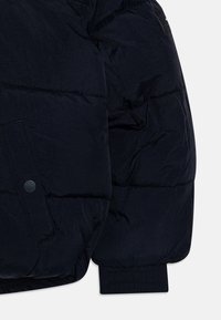 Tmavě modrá vatovaná bunda s prošívaným vzorem, elastickými manžetami, velkou nášivkou s knoflíkem a hladkou, lesklou texturou.