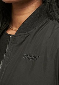 Kvinne i en svart jakke med ribbet krage og "snipes" brodert på brystet, lagdelt over en svart topp og med et gullkjede.
