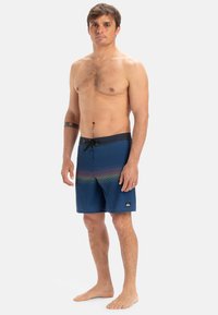 Shorts de bain bleus avec un motif de couleur dégradée, tissu texturé et une taille noire avec cordon de serrage. Logo visible sur la jambe inférieure.