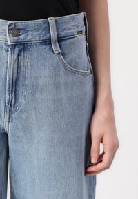 Light blue denim jeans with a straight cut, featuring a silver button and a small black label on the waistband. Textured fabric with visible stitching.