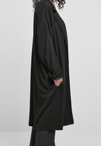 Cardigan lungo oversize nero, realizzato in tessuto morbido, con colletto a risvolto, tasche laterali e maniche arrotolate. Design semplice senza motivi.