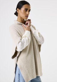 Woman with dark hair in a low bun wearing a beige wool poncho over a white long-sleeve blouse with lace sleeves and pale blue jeans.