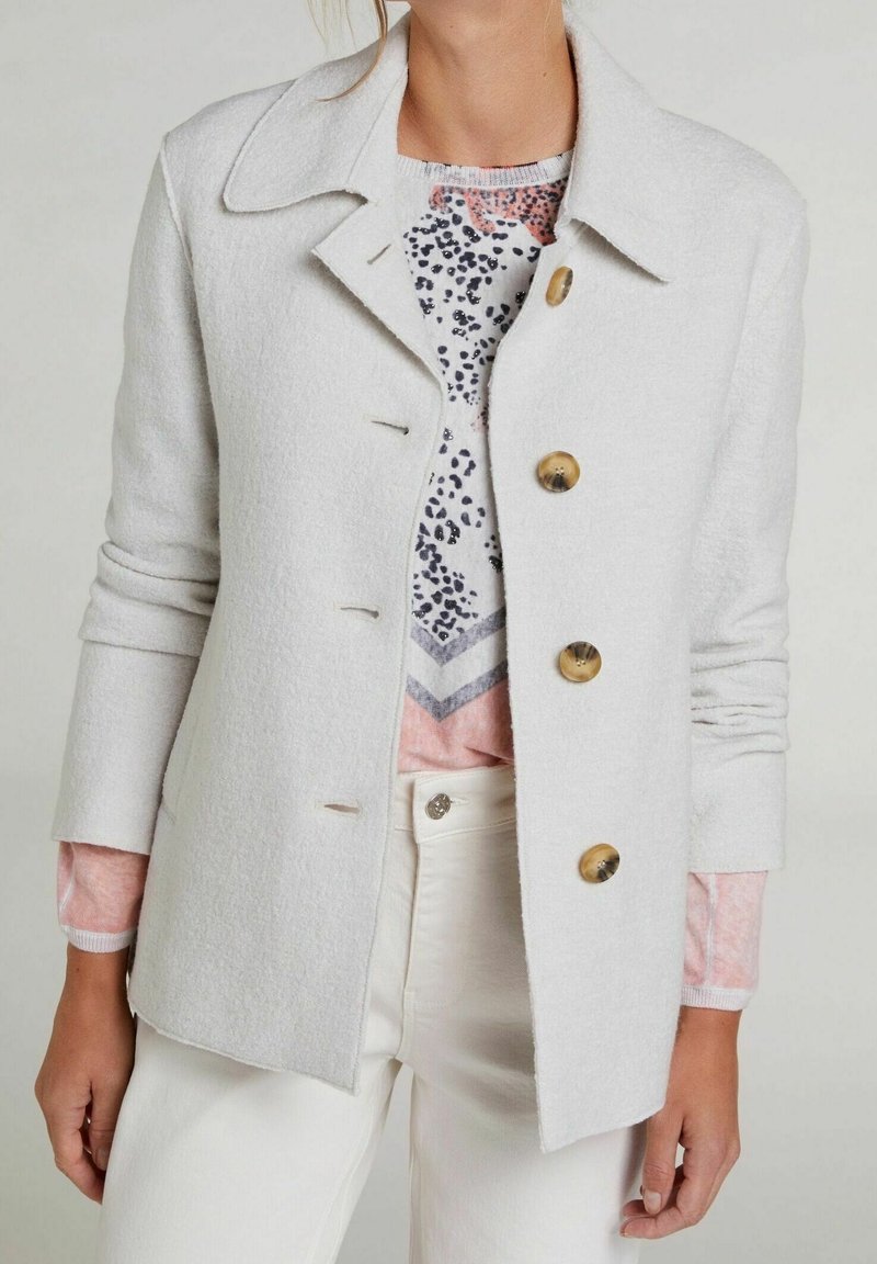 Femme portant un manteau boutonné gris clair sur un haut rose à motifs avec un pantalon blanc sur un fond uni.
